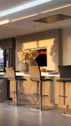 A modern kitchen interior with three sleek bar stools lined up against a light wooden counter. Above the counter, there is a large decorative vase filled with cream-colored flowers. Soft, natural light casts shadows on the walls and floor, creating a warm and inviting atmosphere. A flat screen TV is mounted on the wall behind the counter.