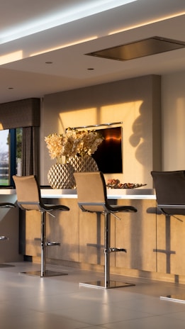 A modern kitchen interior with three sleek bar stools lined up against a light wooden counter. Above the counter, there is a large decorative vase filled with cream-colored flowers. Soft, natural light casts shadows on the walls and floor, creating a warm and inviting atmosphere. A flat screen TV is mounted on the wall behind the counter.