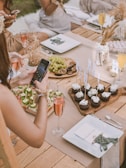 A rustic outdoor dining setup on a wooden table with elegant place settings and assorted refreshments. People are seated, with a hand holding a phone to take a photo of the arrangement of appetizers and cupcakes adorned with flags. A platter of grapes and figs is nearby, along with glasses of rosé and champagne.
