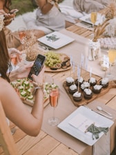 A rustic outdoor dining setup on a wooden table with elegant place settings and assorted refreshments. People are seated, with a hand holding a phone to take a photo of the arrangement of appetizers and cupcakes adorned with flags. A platter of grapes and figs is nearby, along with glasses of rosé and champagne.