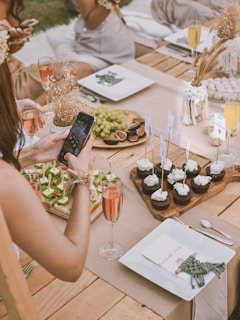 A rustic outdoor dining setup on a wooden table with elegant place settings and assorted refreshments. People are seated, with a hand holding a phone to take a photo of the arrangement of appetizers and cupcakes adorned with flags. A platter of grapes and figs is nearby, along with glasses of rosé and champagne.