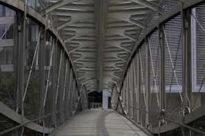 A modern, enclosed pedestrian bridge with metal and glass construction stretches across an urban area. The symmetrical design features intricate steel beams and support structures. The walkway has a partially transparent floor and an arched roof.