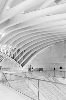 A modern architectural interior with sweeping, curved beams creating an open and spacious atmosphere. The area is dominated by sleek staircases and transparent glass railings, all in a monochromatic scheme. The design is minimalistic and emphasizes clean lines and geometric shapes.