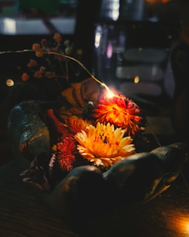 A collection of vibrant flowers, illuminated by a small string light, rests gently in a stone or metallic hand sculpture. The flowers are predominately in shades of yellow, orange, and red, surrounded by darker foliage. The surrounding environment is dimly lit, creating a warm and cozy ambiance.