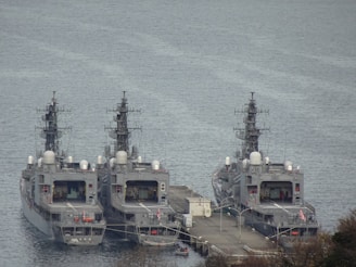 Three navy ships are docked side by side at a pier on a calm body of water. The ships are grey in color and appear to be equipped with various communication and radar equipment mounted on top. The pier has a few structures and pathways connecting the ships. There are also lifeboats and other small boats visible near the pier. The surrounding area includes some vegetation and trees.