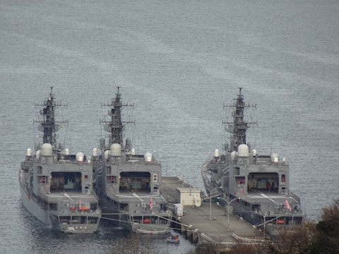 Three navy ships are docked side by side at a pier on a calm body of water. The ships are grey in color and appear to be equipped with various communication and radar equipment mounted on top. The pier has a few structures and pathways connecting the ships. There are also lifeboats and other small boats visible near the pier. The surrounding area includes some vegetation and trees.