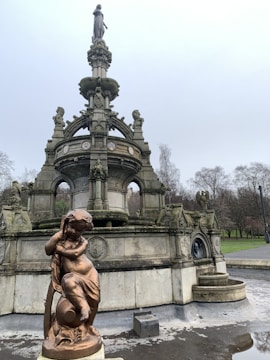 A detailed stone monument with intricate carvings, featuring a prominent statue of a person at the top. Below, a bronze statue of a child is seen leaning against an orb. The monument is surrounded by a park environment with trees in the background under a cloudy sky.