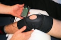 A person is applying a black knee brace to another person's leg, which is resting on a pillow. The person is also holding a handheld electronic device with a small screen displaying some controls and information, likely related to therapy or treatment.
