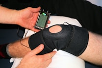 A person is applying a black knee brace to another person's leg, which is resting on a pillow. The person is also holding a handheld electronic device with a small screen displaying some controls and information, likely related to therapy or treatment.