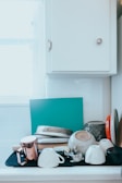 A collection of kitchen items are stacked on a countertop against a white tile backsplash. Several cups are upside down, and a metal teapot or mug with a copper finish is visible. Plates and a teal cutting board are leaning against the wall, and there is a white cabinet above them.