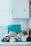 A collection of kitchen items are stacked on a countertop against a white tile backsplash. Several cups are upside down, and a metal teapot or mug with a copper finish is visible. Plates and a teal cutting board are leaning against the wall, and there is a white cabinet above them.