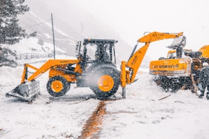 Heavy machinery operates in a snowy environment with a yellow excavator and a loader clearing the snow. Snowflakes are visibly falling, and the area is surrounded by snow-covered mountains and trees.