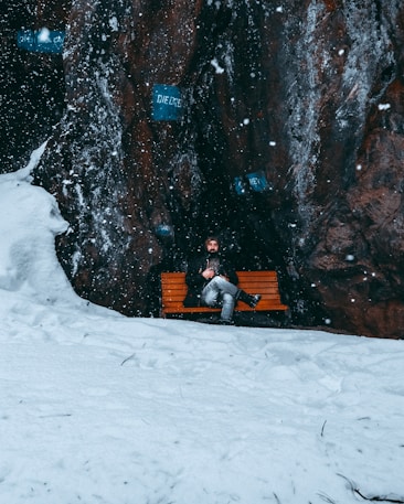 A person wearing a winter coat is sitting on a wooden bench amidst a snowy landscape. The scene includes a rugged rock wall in the background with visible snowflakes falling. Various blue signs with text are embedded into the rock surface, creating an intriguing contrast against the natural elements.