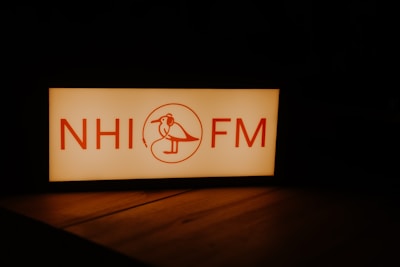 A rectangular sign with the letters 'NHI FM' and a stylized bird wearing headphones in between. The text and illustration are in a warm red color against a glowing white background. The surrounding environment is dark, highlighting the illuminated sign.