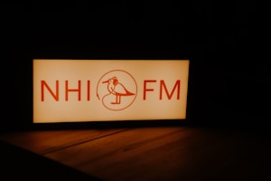 A rectangular sign with the letters 'NHI FM' and a stylized bird wearing headphones in between. The text and illustration are in a warm red color against a glowing white background. The surrounding environment is dark, highlighting the illuminated sign.