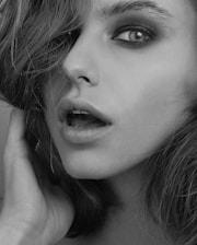 A close-up black and white portrait of a person's face. The image highlights the left eye, partially open lips, and nostrils. Hair is slightly tousled, and a hand is gently touching the cheek. The makeup is subtle, with emphasis on the eye.