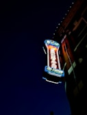 A neon sign is mounted on the side of a building, illuminated against a dark night sky. The sign features vibrant colors, including red, blue, and green, with intricate detailing and lettering that likely indicates a business name or type.