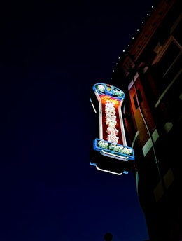 A neon sign is mounted on the side of a building, illuminated against a dark night sky. The sign features vibrant colors, including red, blue, and green, with intricate detailing and lettering that likely indicates a business name or type.