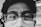 A close-up black and white photograph of a person's face, focusing on their eyes behind thick-rimmed glasses. The person is wearing a surgical mask that partially covers the face. The lighting casts soft shadows, emphasizing the reflection in the lenses and creating a contemplative mood.