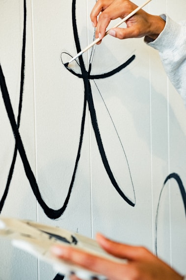 A person is painting large black calligraphic strokes on a light-colored wall. The hand holding the brush is steady and focused on creating smooth, flowing lines, while the other hand appears to be holding a palette.