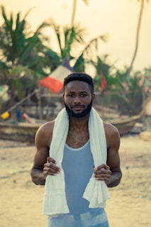 A man stands on a beach with a white towel draped over his shoulders. He wears a light-colored tank top. In the background, palm trees sway gently, and a boat with colorful flags can be seen, suggesting a coastal, possibly tropical setting.