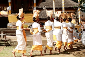 A group of women wearing traditional Balinese attire, featuring white garments with golden yellow sashes and elaborate headpieces, walk in a line, possibly participating in a cultural ceremony or dance. The background shows a traditional Balinese temple with intricate carvings and decorations.