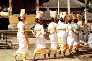 A group of women wearing traditional Balinese attire, featuring white garments with golden yellow sashes and elaborate headpieces, walk in a line, possibly participating in a cultural ceremony or dance. The background shows a traditional Balinese temple with intricate carvings and decorations.
