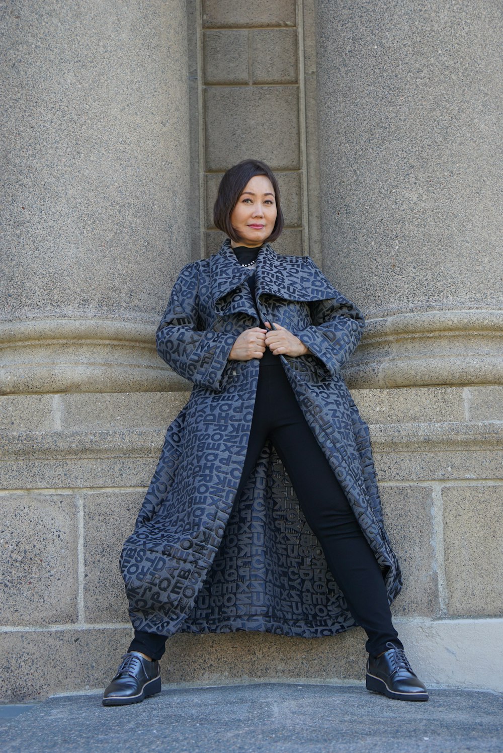 灰色のコンクリートの壁のそばに立つ灰色のコートを着た女性