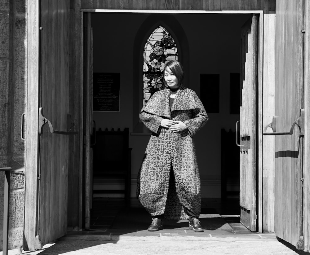 grayscale photo of man in coat standing in front of door