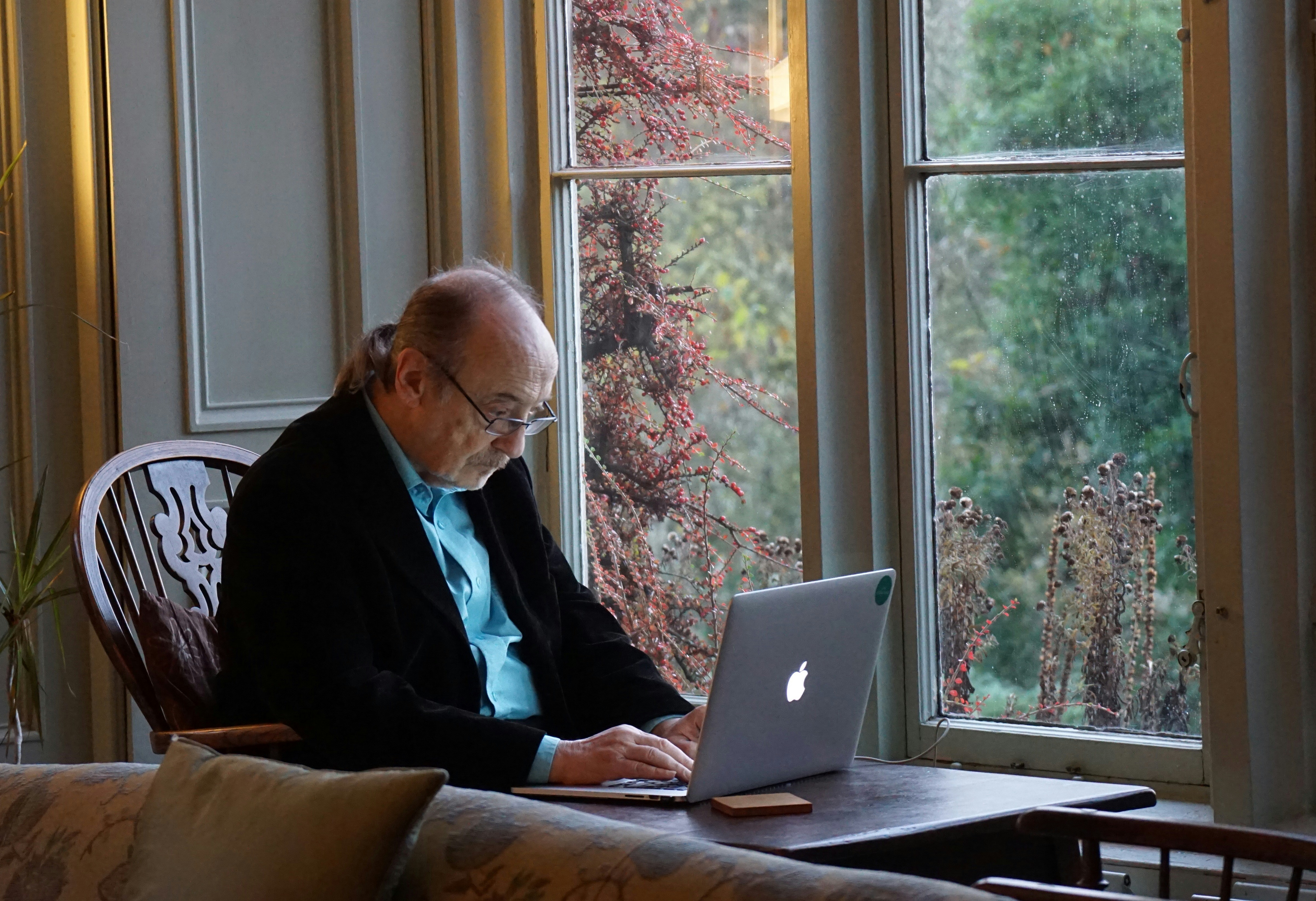 Elderly man sitting on a chair using a laptop on a table inside a house.