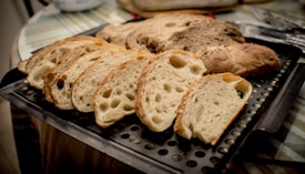 A loaf of bread cut into several slices, placed on a perforated metal tray. The bread has a rustic appearance with a light brown crust and a soft, airy interior with visible air pockets. The background includes part of a tabletop with a plaid pattern and some cutlery.
