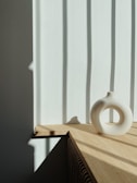 A minimalist ceramic vase with a circular design sits on a light wooden surface. The scene is bathed in soft natural light, creating shadows from nearby vertical blinds on a white wall.