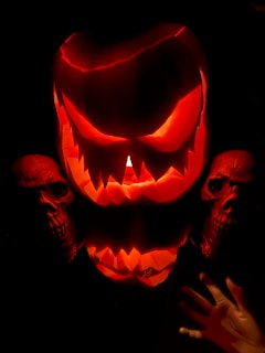 A large, menacingly carved pumpkin glows with an intense orange light against a dark background. The pumpkin's expression is eerie, with sharply pointed teeth and slanted eyes. Flanking the pumpkin are two human skulls, adding to the sinister ambiance. A hand, likely part of a Halloween decoration, reaches out from the lower right.