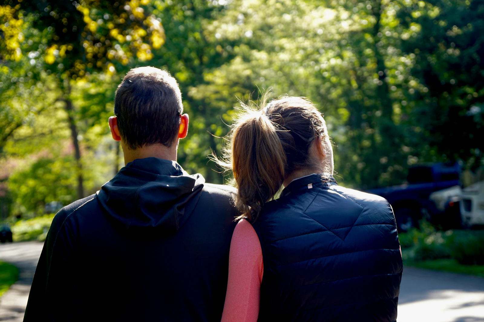 Two people walking side by side along a tree-lined path, seen from behind.
