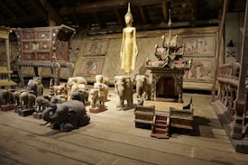 A wooden room filled with a variety of carved wooden sculptures, including elephants and a golden statue. The sculptures are intricately detailed, and some resemble shrines or altars. The background features woven bamboo panels and other large furniture pieces, creating a rustic and cultural atmosphere.