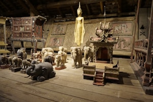 A wooden room filled with a variety of carved wooden sculptures, including elephants and a golden statue. The sculptures are intricately detailed, and some resemble shrines or altars. The background features woven bamboo panels and other large furniture pieces, creating a rustic and cultural atmosphere.