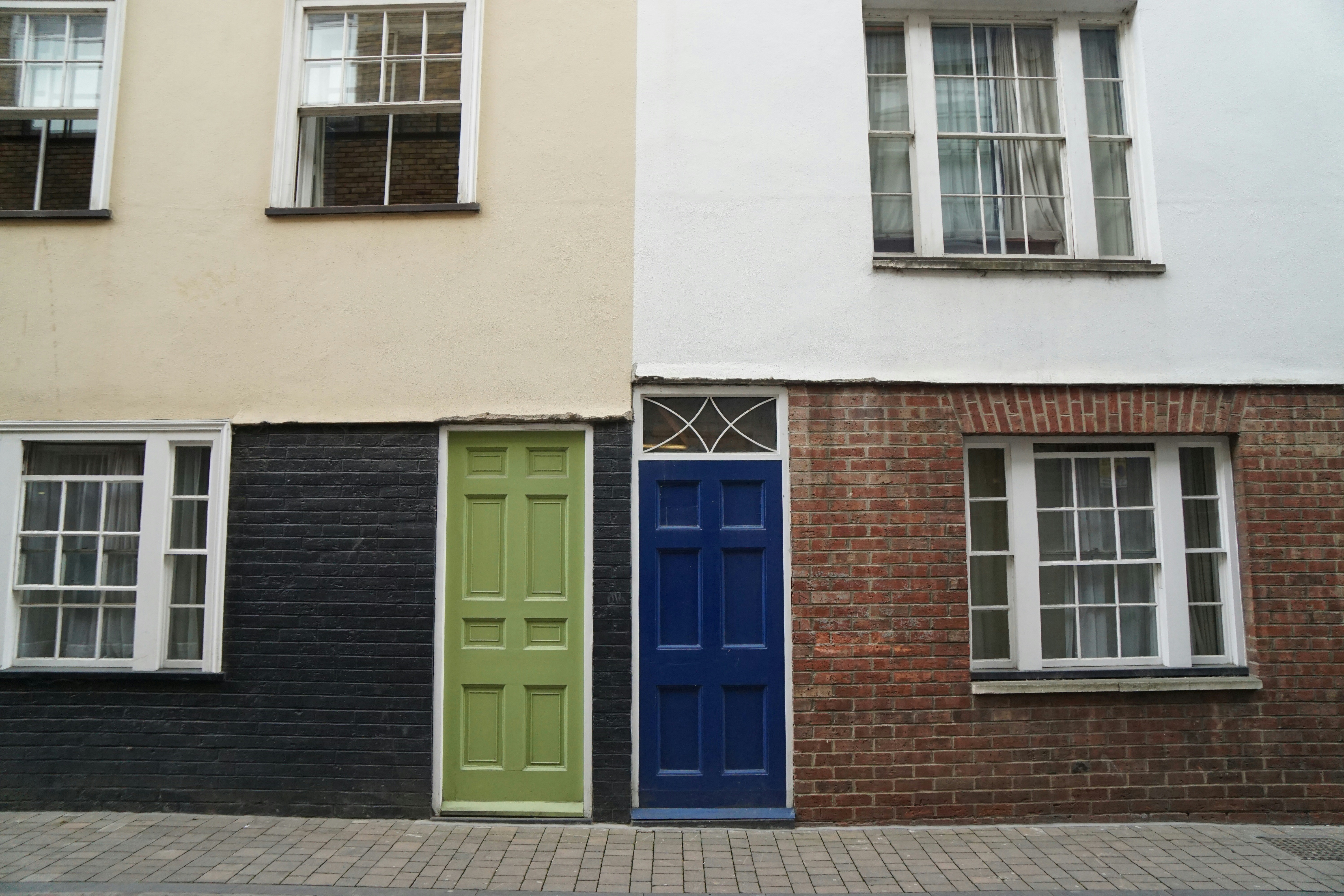 A street-level view of a building facade featuring two doors and three windows. The left side of the building has a green door with a black brick wall and a large window with white trim above and to the side. The right side of the building features a blue door with a red brick wall, flanked by a similar style window. The upper portion of the building is painted white, and the street consists of cobblestones.