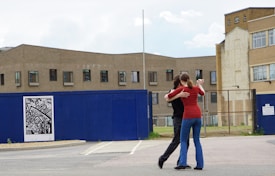 Two people are embracing in close proximity in an outdoor setting, possibly dancing on an open area with buildings in the background. A large blue fence with an abstract black and white poster attached is visible, along with several windows and a paved surface.