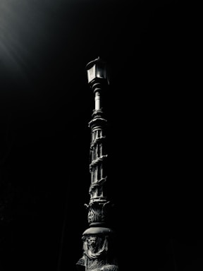 A vintage-style street lamp with ornate details stands against a dark background, its design featuring intricate patterns and curves. The lighting highlights the metallic texture of the lamp post.