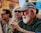 An elderly man with a white beard and glasses is wearing a grey cap and a dark green shirt, sitting outdoors. He appears to be clapping or rubbing his hands. In the blurred background, two other people are visible; one is wearing sunglasses and has blonde hair.