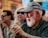 An elderly man with a white beard and glasses is wearing a grey cap and a dark green shirt, sitting outdoors. He appears to be clapping or rubbing his hands. In the blurred background, two other people are visible; one is wearing sunglasses and has blonde hair.