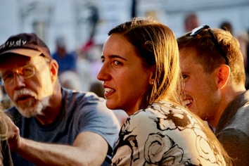 A group of three people engaged in conversation, with warm sunlight highlighting their faces. The man on the left is wearing a cap and glasses, the woman in the center has long brown hair and a floral patterned shirt, and the man on the right has short hair and sunglasses perched on his head.