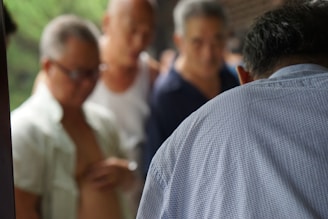 A group of elderly men is gathered in a close setting, with one in the foreground wearing a white shirt facing away from the camera. The background is slightly blurred, focusing more on the individuals' expressions and interactions.