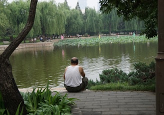 man in white shirt sitting on concrete pavement near lake during daytime