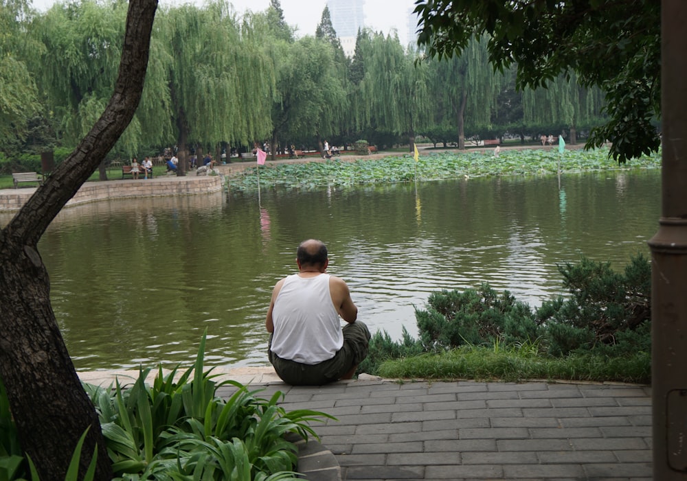 Mann im weißen Hemd sitzt tagsüber auf Betonpflaster in der Nähe des Sees
