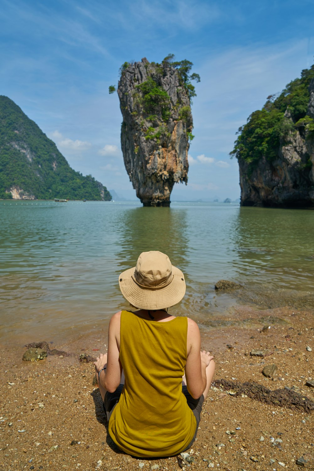 man in yellow tank top and brown fedora hat sitting on brown rock near body of near near near near