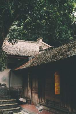 A traditional wooden house with a tiled roof surrounded by dense greenery. The structure has a rustic charm, with wooden doors and a small staircase leading to the entrance. Light filters through the trees above, creating a serene atmosphere.