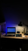 A dimly lit desk with a laptop displaying a webpage. The desk is organized with a few books on the side, a mouse resting on a mouse pad, and a lamp casting light onto the workspace. A figurine and a small speaker are also present, adding personal touches to the setup.
