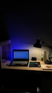 A dimly lit desk with a laptop displaying a webpage. The desk is organized with a few books on the side, a mouse resting on a mouse pad, and a lamp casting light onto the workspace. A figurine and a small speaker are also present, adding personal touches to the setup.
