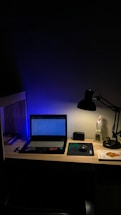 A dimly lit desk with a laptop displaying a webpage. The desk is organized with a few books on the side, a mouse resting on a mouse pad, and a lamp casting light onto the workspace. A figurine and a small speaker are also present, adding personal touches to the setup.